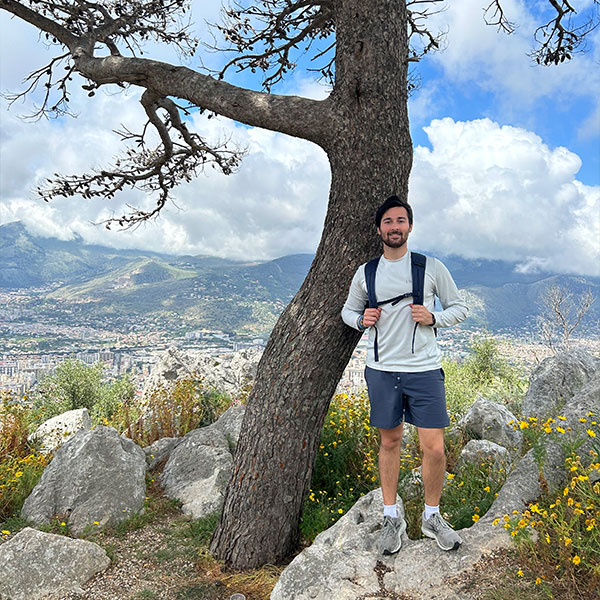 a male student wearing a backpack stands on white rocks next to a tree