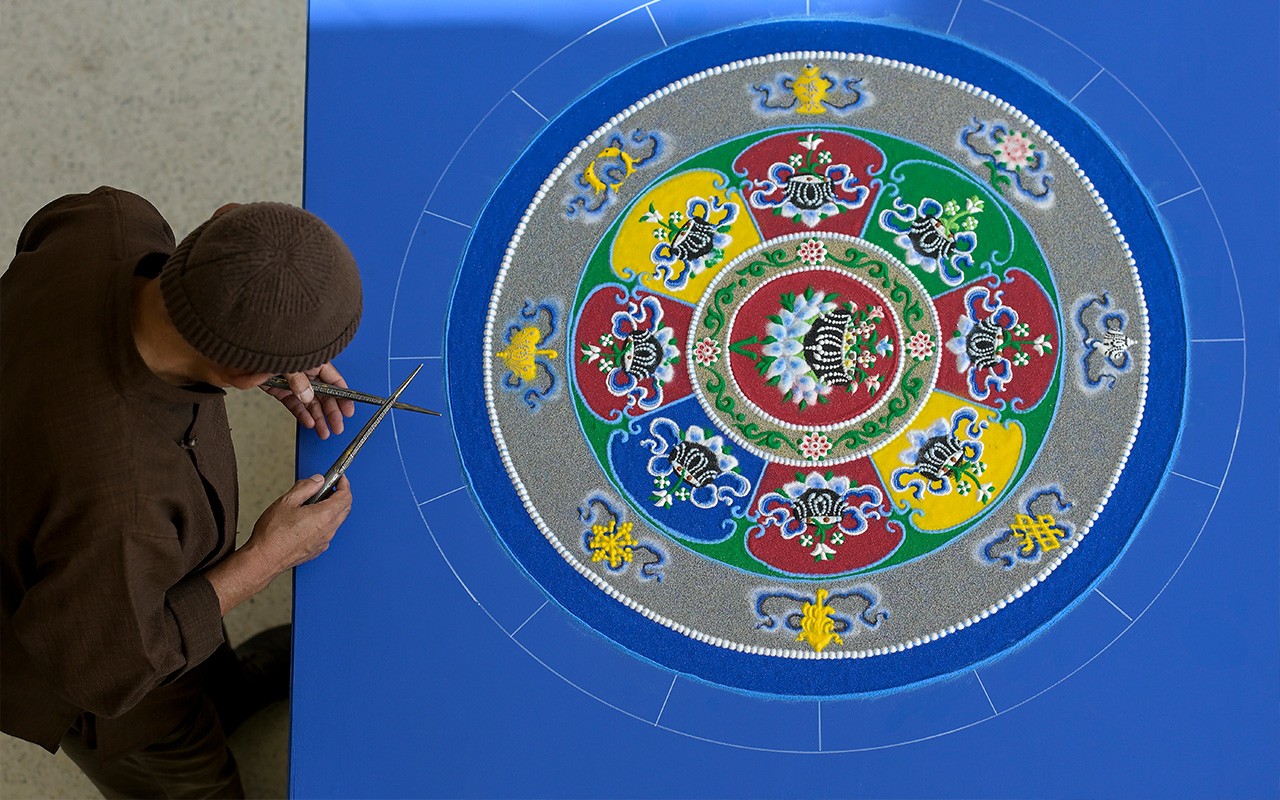 A Tibetan scholar wearing a brown cap and robe holds the tools he's using to create an intricate sand mandala. 