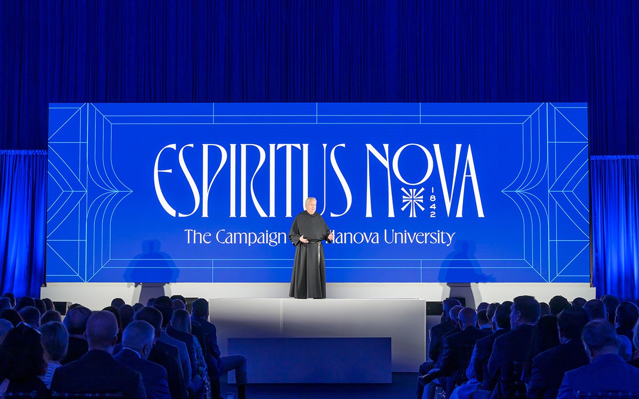 Father Peter wearing his black habit stands before a large Espiritus Nova banner while speaking to an audience of Villanovans.