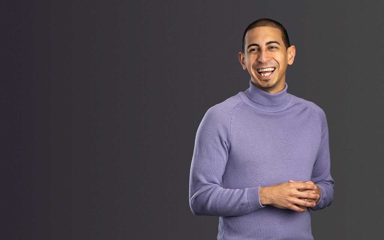 Associate Professor Alexander Diaz-Lopez stands smiling with his hands clasped, wearing a lavender turtleneck and black pants.